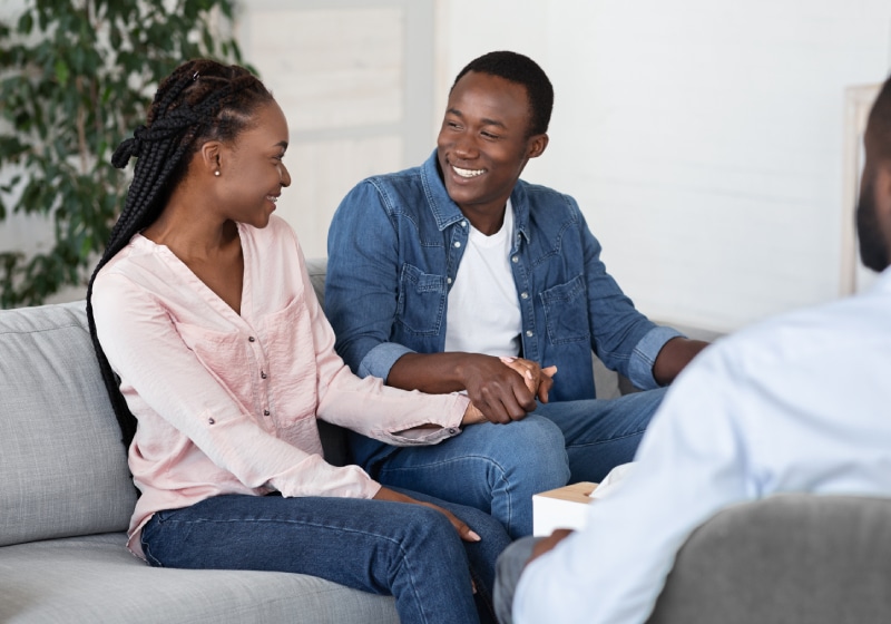 casal negro fazendo entrevista sentados em sofá um olhando para outro sorridentes 