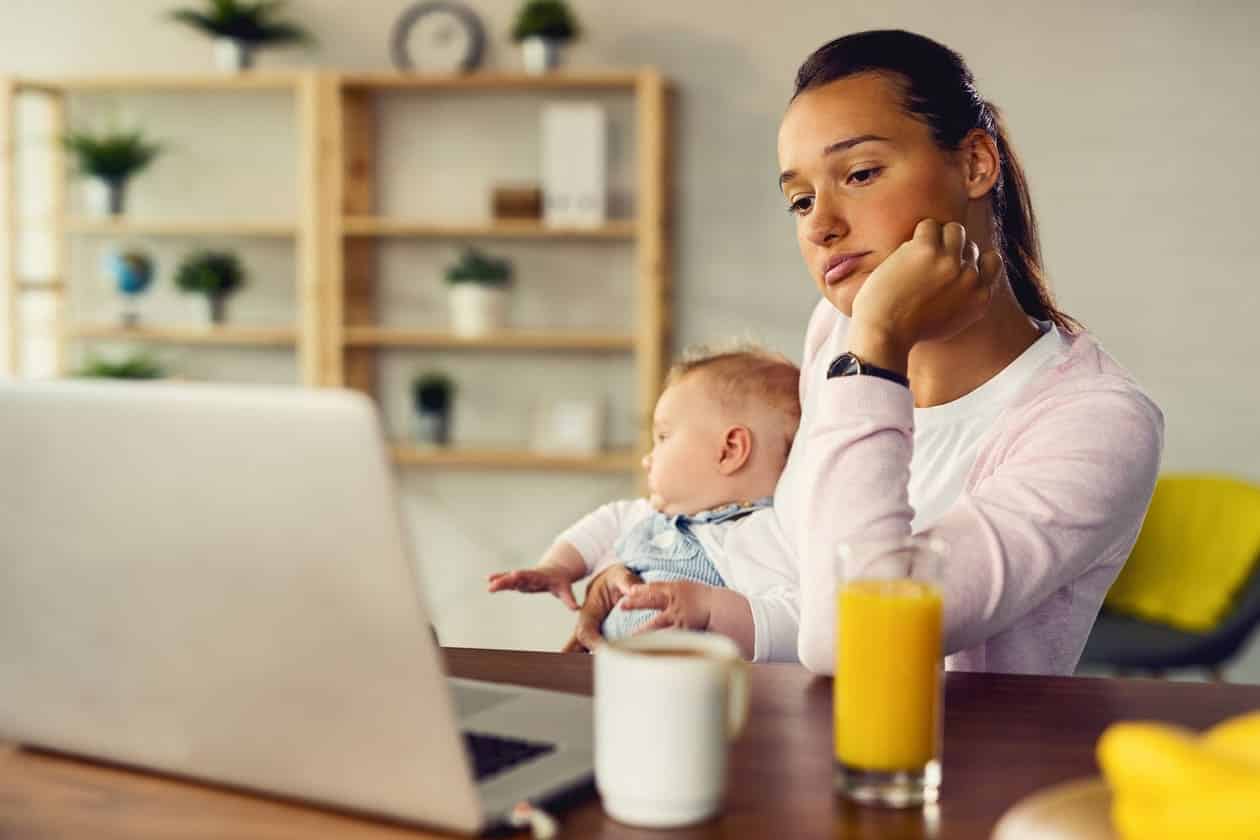 mulher olha pensativa para o computador enquanto segura o bebê no colo