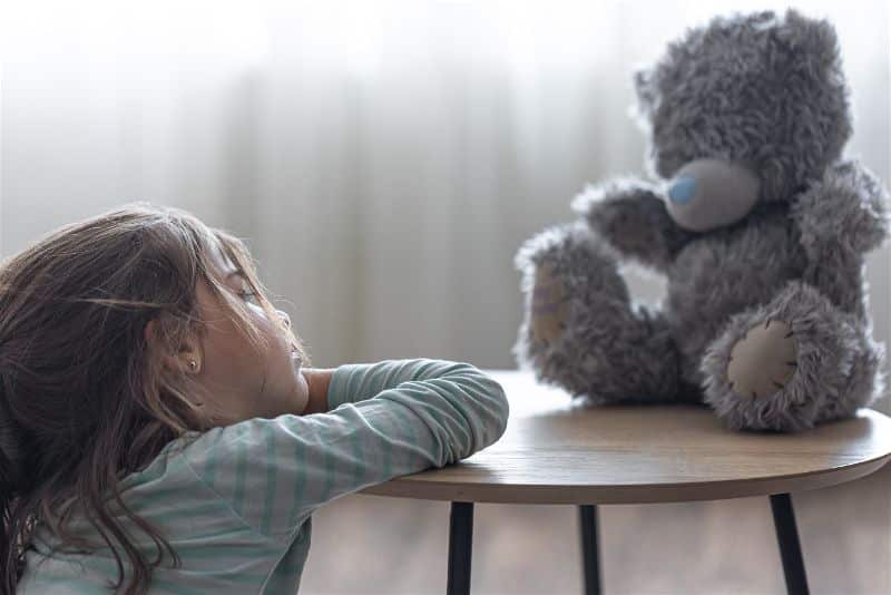 criança apoiando a cabeça com a mão sentada de frente para um urso de pelúcia