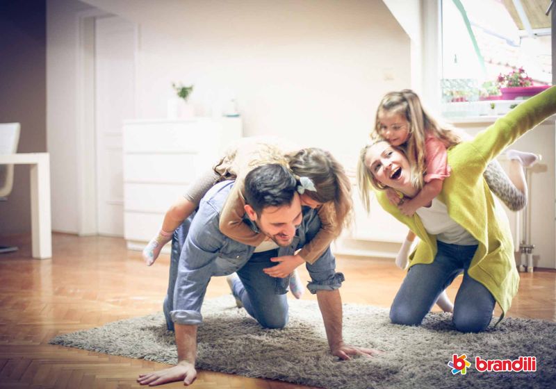 Pai, mãe e duas filhas brincam de cavalinho em cima do tapete.