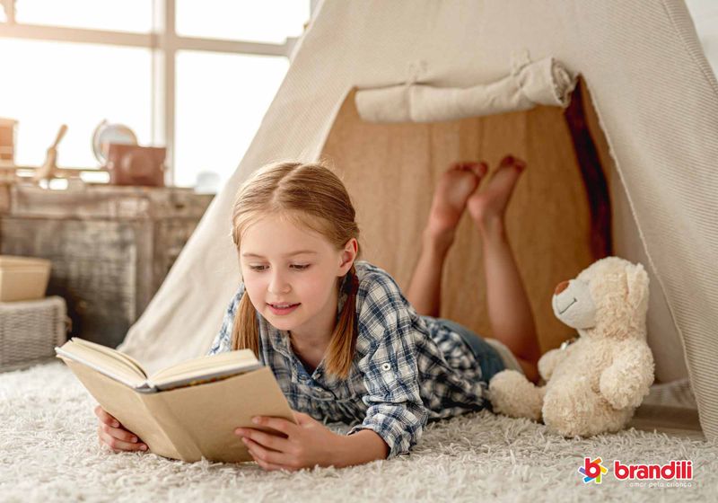 Menina loira com pijama xadrez lê livro sorrindo dentro de sua cabaninha.