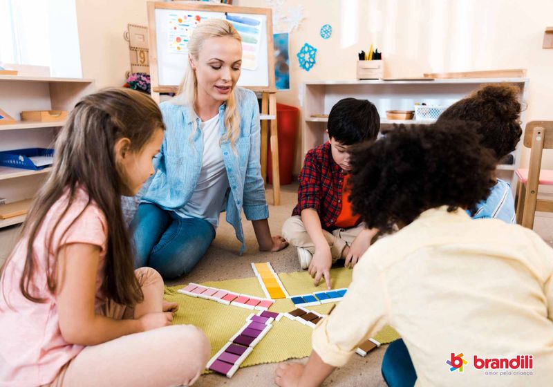 Professora e quatro alunos analisam atividade com cores no chão da sala de aula.