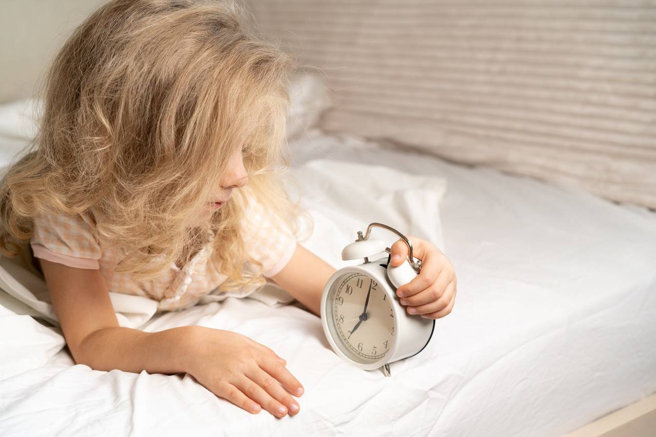 menina pequena deitada na cama segurando um relógio enquanto vê o horário de dormir