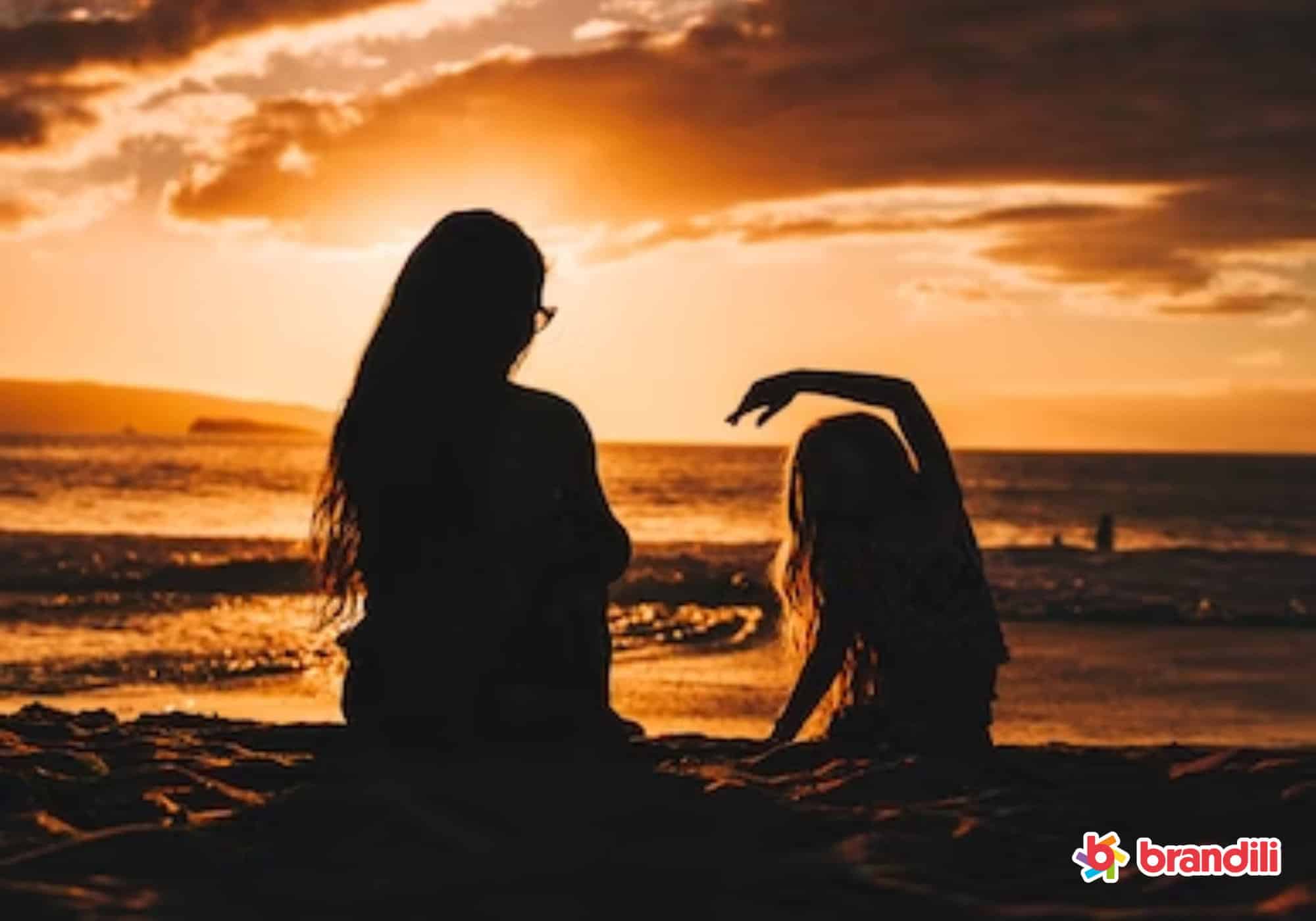 Fotografia contra o sol de mãe e menina de costas sentadas se divertindo na praia