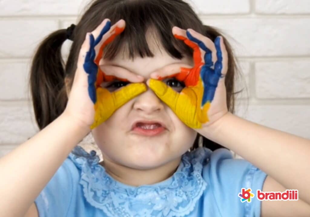 Criança menina faz um óculos com as mãos pintadas de tinta