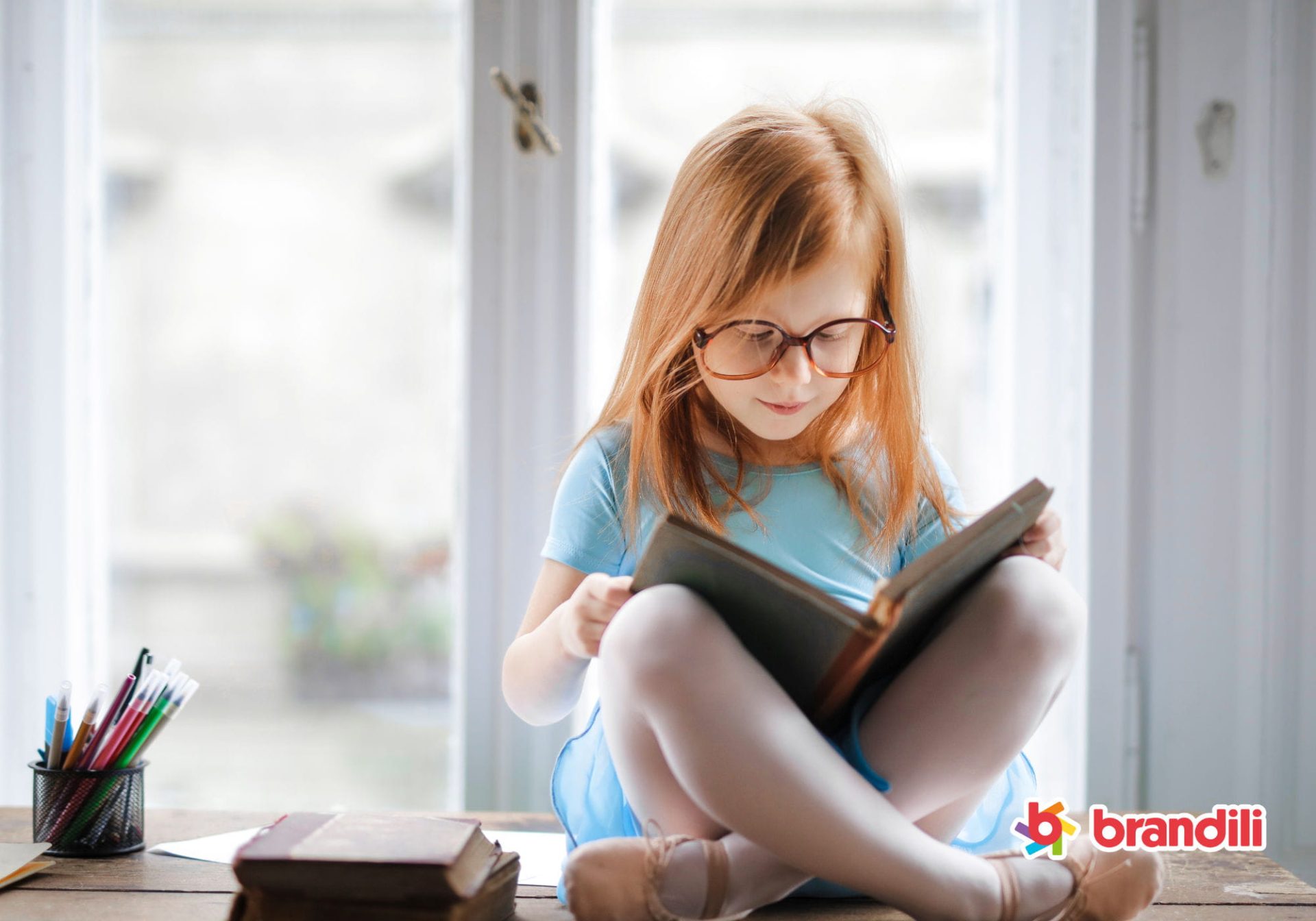 Criança de óculos sentada próxima à janela lendo um livro.