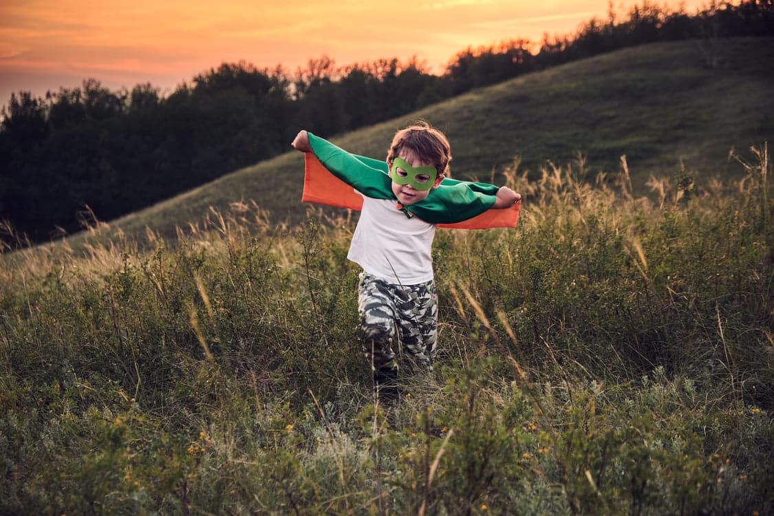 menino com uma capa verde e uma roupa temática de super heroi correndo em um campo aberto