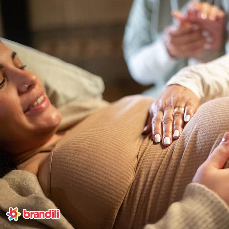 mulher gravida sorridente com roupas casuais deitada na cama segurando a mao na barriga