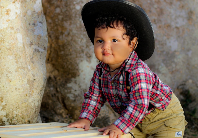 bebe se apoiando em caixote com look de cawboy camisa xadrez calça bege e chapéu de cawboy 