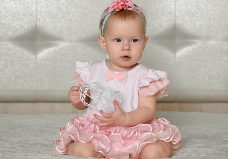 bebê sentado na cama com vestido rosa e branco com brinquedinho na mão 