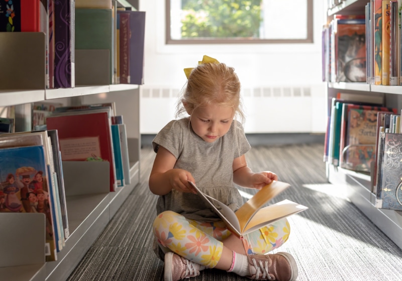 garotinha loira sentada no chão lendo um livro em biblioteca 