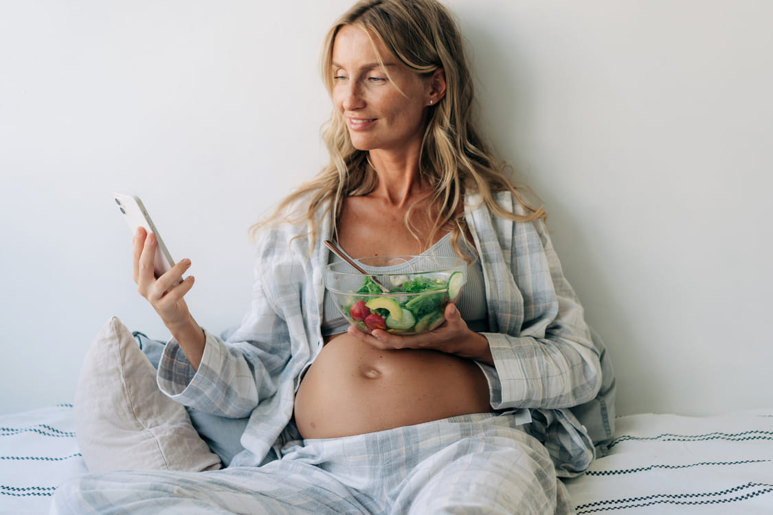 mulher grávida olha para o celular enquanto come um prato de salada
