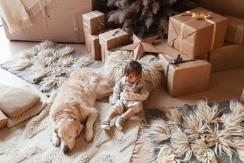 Criança sentada em sala com vários tapetes nas cores bege e cinza, vários presentes embalados ao lado, com cachorro dormindo próximo da criança