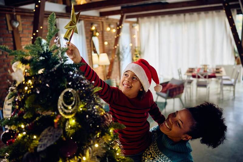 Criança sendo segurada no colo, e colocando estrela decorativa no topo da árvore natalina 