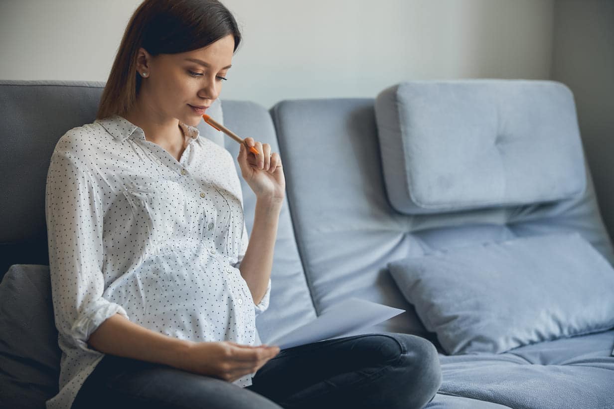 grávida sentada no sofá com cara pensativa e um papel e caneta na mão