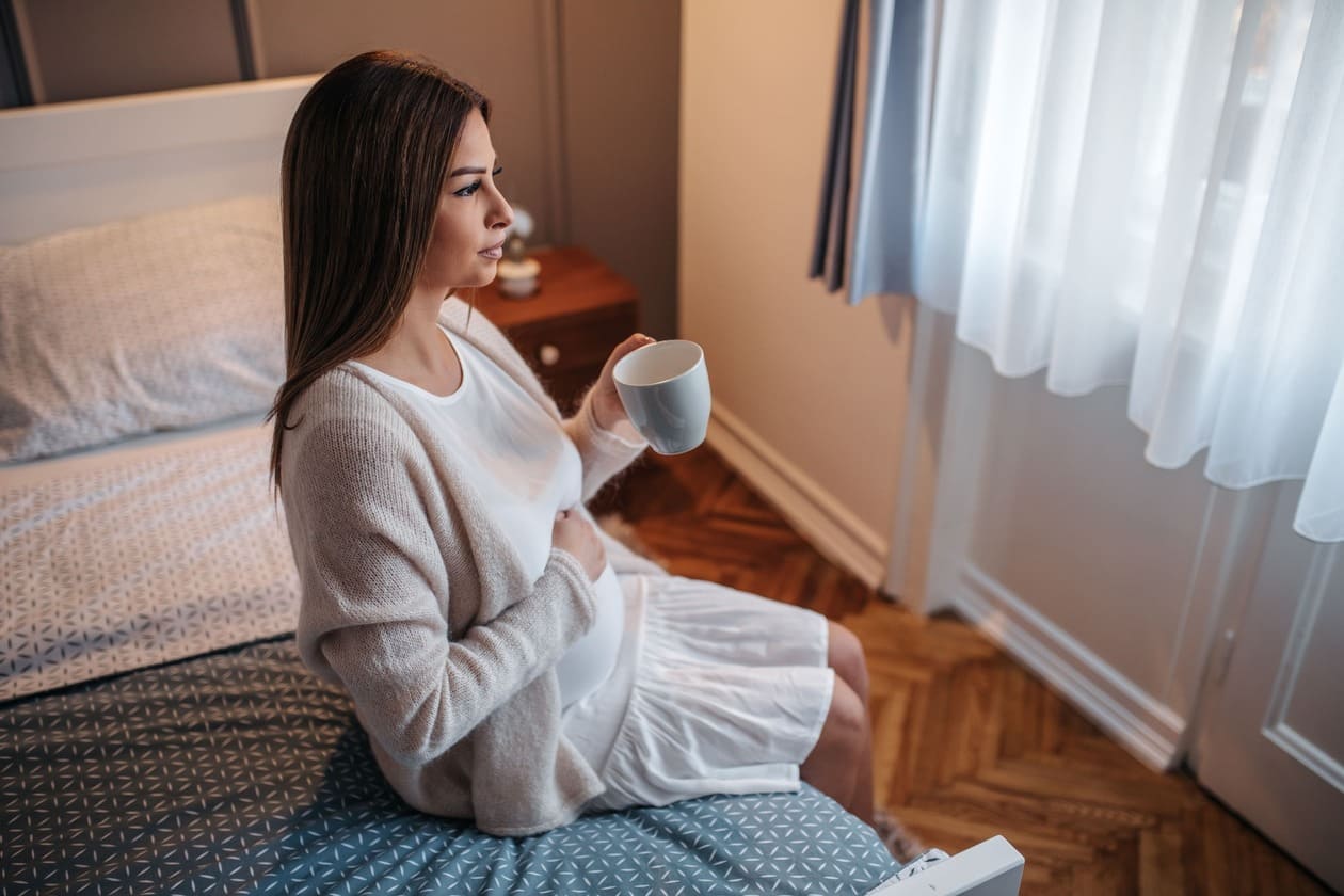 grávida de 9 semanas sentada na beira da cama, olhando para a janela e tomando chá