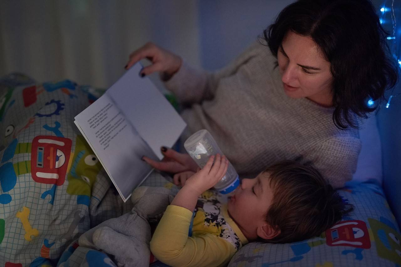 mãe lendo as Aventuras de Alice no País das Maravilhas para seu filho enquanto ele toma leite na mamadeira para dormir