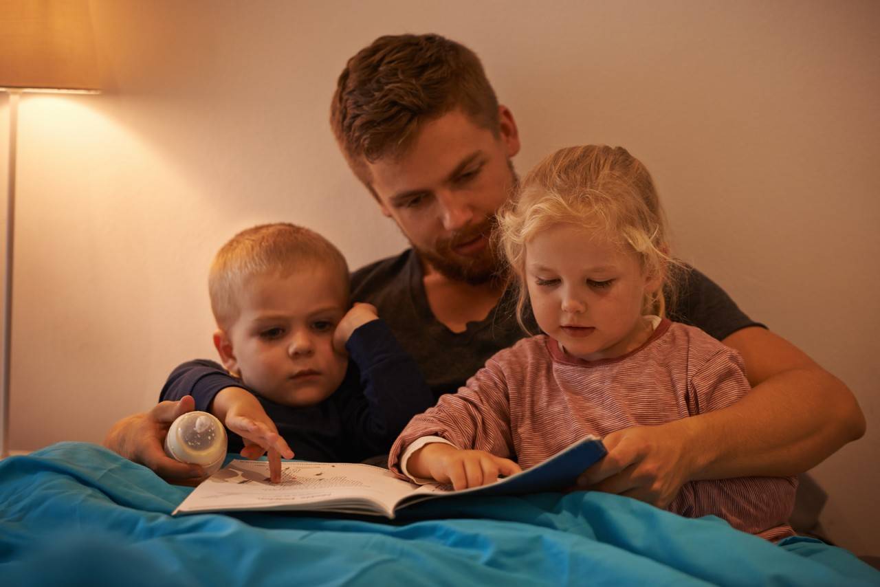 pai sentado na cama com os dois filhos lendo histórias para eles dormirem