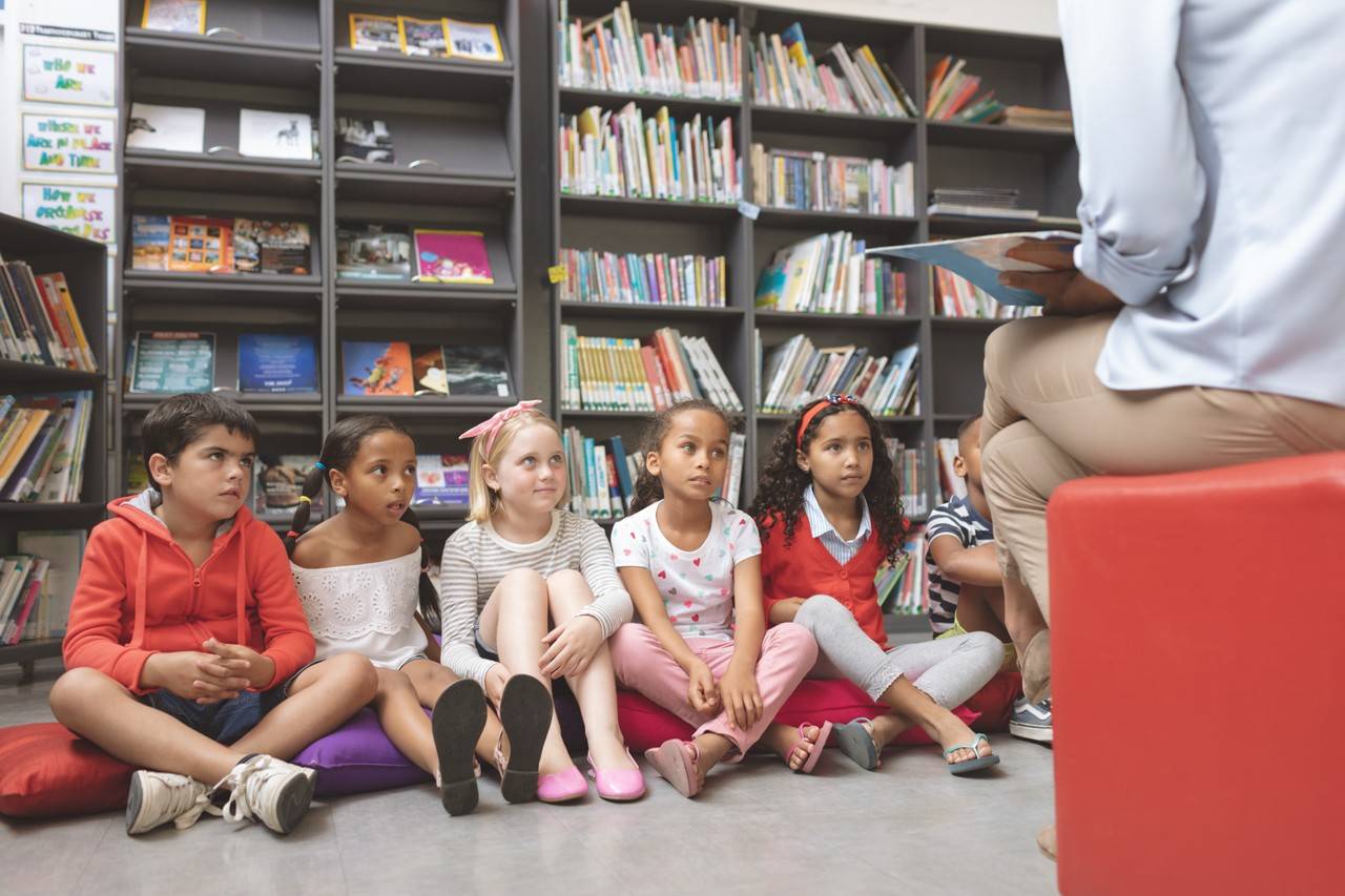 crianças sentadas em frente de uma professora que está lendo um livro para os mais novos