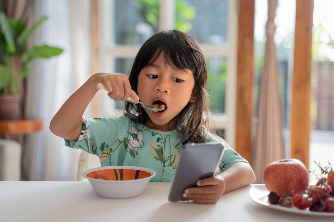 criança comendo comida enquanto usa o celular