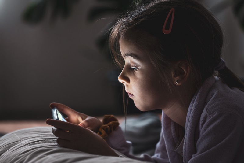 menina pequena deitada na cama jogando no celular com fone de ouvido